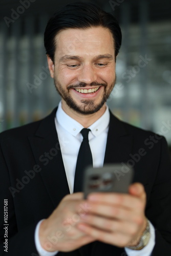 A man in a suit is smiling while looking at his cell phone. He is wearing a tie and he is in a professional setting