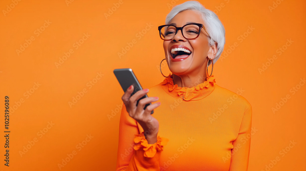 Smiling elderly woman in an orange outfit laughing while using a smartphone against an orange background. Smiling elderly woman in an orange outfit laughing while using a smartphone against an orange background.
