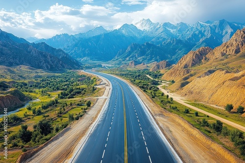 A long, straight highway cuts through a mountain valley, with green fields and a river on either side. The road leads towards snow-capped peaks in the distance, under a blue sky with white clouds.