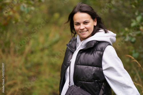 Photography Young beautiful smiling dark-haired girl in vest and hoodie standing against blurred forest background