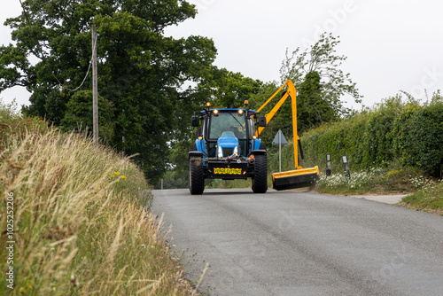 Blue Verge Cutting Agricultural Tractor with Yellow Grass Cutting Arm Trimming Grass Beside Rural Country Road with Farm Hedgerow