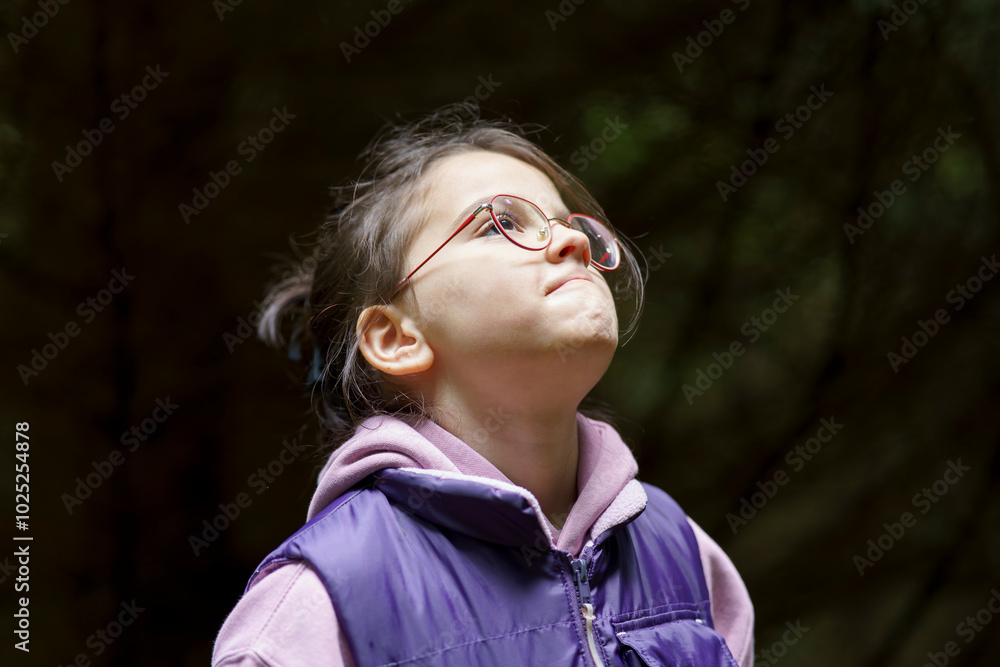 Little beautiful girl with glasses in a hoodie and vest against the background of a dark autumn forest. The concept of a happy childhood in nature Little beautiful girl with glasses in a hoodie and vest against the background of a dark autumn forest. The concept of a happy childhood in nature