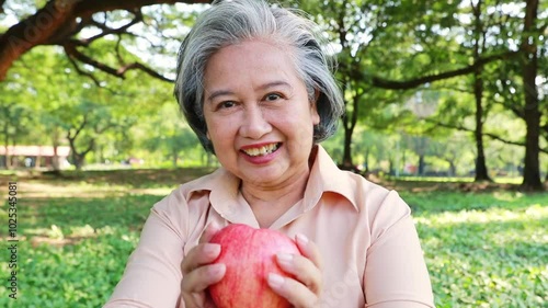 Senior Asian woman camping in a park shows her desire to eat sweet juicy and sour red apple with a smiling face : Elderly women take care of their health and like to eat fruit.