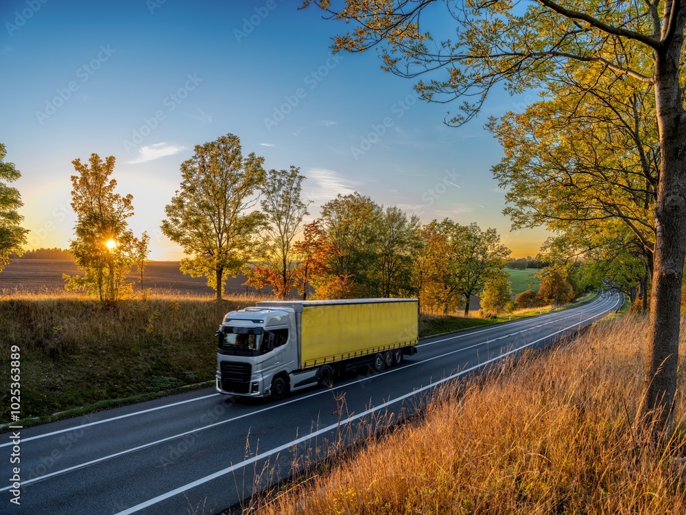 Silver truck driving on an asphalt road between rows of leafy trees in autumn colors in a rural landscape at sunset