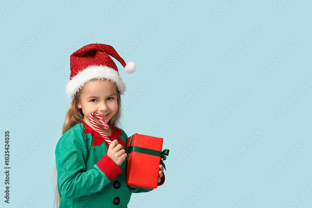 Cute little girl in elf costume with gift box and candy cane on blue background Cute little girl in elf costume with gift box and candy cane on blue background