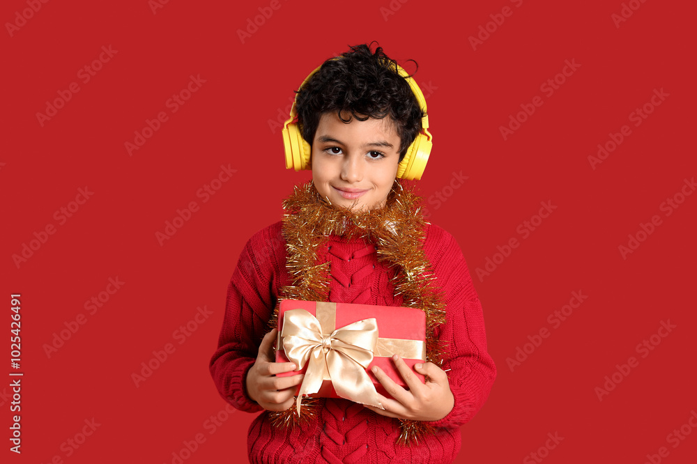Cute little boy in headphones with tinsel and Christmas gift box on red background Cute little boy in headphones with tinsel and Christmas gift box on red background