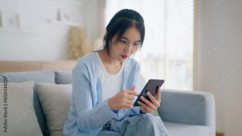 Smiling pretty young Asian woman sitting on cozy couch, using funny mobile apps in living room. Woman at home, Doing online shopping, Messaging friends, Posting on social media Smiling pretty young Asian woman sitting on cozy couch, using funny mobile apps in living room. Woman at home, Doing online shopping, Messaging friends, Posting on social media