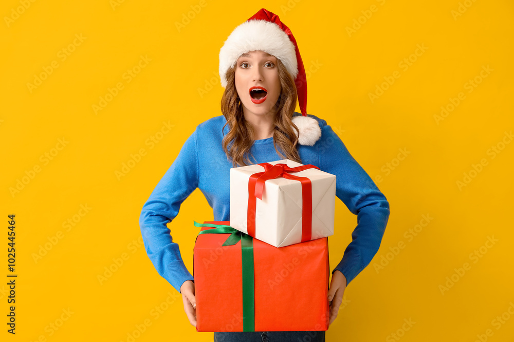 Surprised young woman in Santa hat with Christmas gift boxes on yellow background Surprised young woman in Santa hat with Christmas gift boxes on yellow background