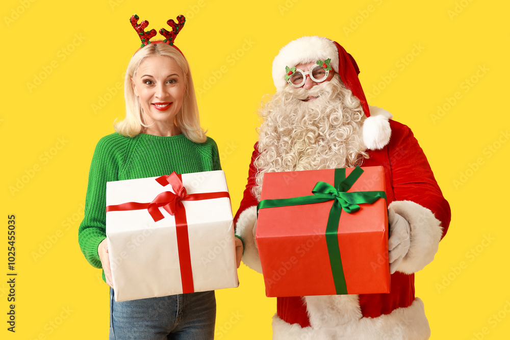 Santa Claus and beautiful mature woman in Christmas deer horns with gift boxes on yellow background Santa Claus and beautiful mature woman in Christmas deer horns with gift boxes on yellow background