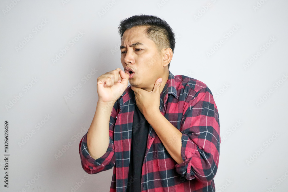 Young Asian man in casual t-shirt coughing into his fist while holding his chest, indicating discomfort or illness. Studio shot on plain background capturing his casual outfit Young Asian man in casual t-shirt coughing into his fist while holding his chest, indicating discomfort or illness. Studio shot on plain background capturing his casual outfit