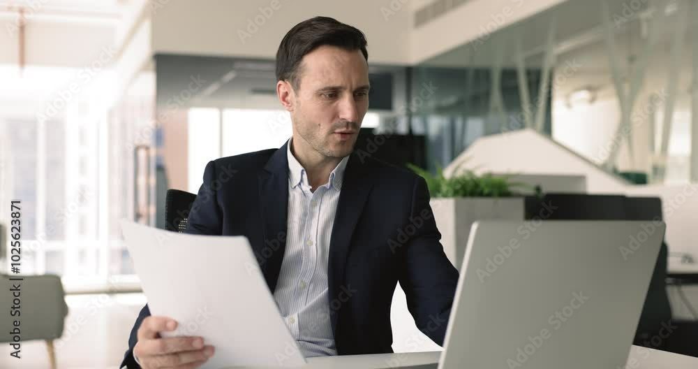Focused businessman sits at desk in office working on laptop, analyze information, learn contract details, agreement conditions, comparing information on computer and in document, make data analysis Focused businessman sits at desk in office working on laptop, analyze information, learn contract details, agreement conditions, comparing information on computer and in document, make data analysis
