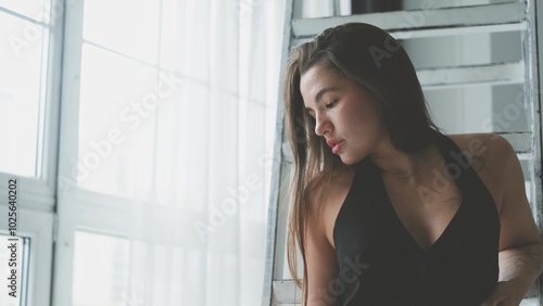 Portrait of young beautiful model posing in black lingerie and stockings near a large window. Calm cute brunette in fashionable lingerie.