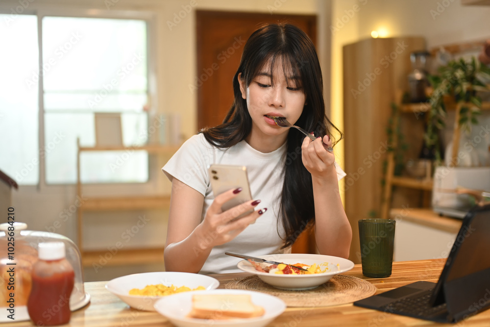 Beautiful young woman enjoying a meal while using her smartphone at a kitchen table Beautiful young woman enjoying a meal while using her smartphone at a kitchen table
