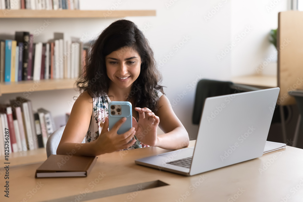 Positive young Indian businesswoman in casual using business app on smartphone for online communication, sitting at laptop in office co-working, workplace, typing work message, smiling Positive young Indian businesswoman in casual using business app on smartphone for online communication, sitting at laptop in office co-working, workplace, typing work message, smiling