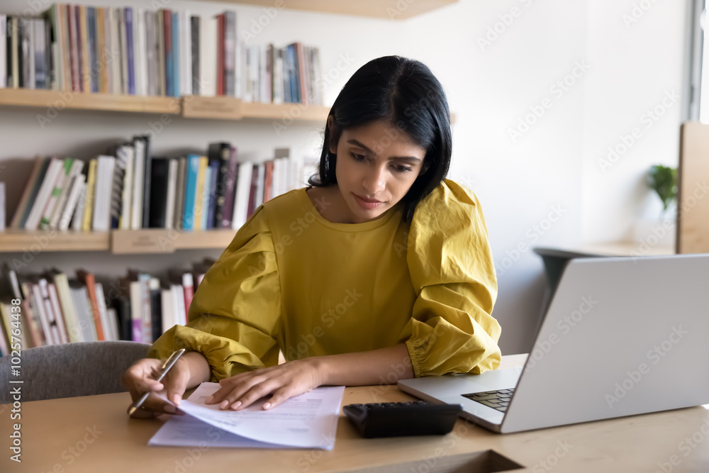 Serious young Indian accountant girl analyzing contract, financial terms, checking investment agreement, using calculator for accounting. Student counting loan for education fees, reading documents Serious young Indian accountant girl analyzing contract, financial terms, checking investment agreement, using calculator for accounting. Student counting loan for education fees, reading documents
