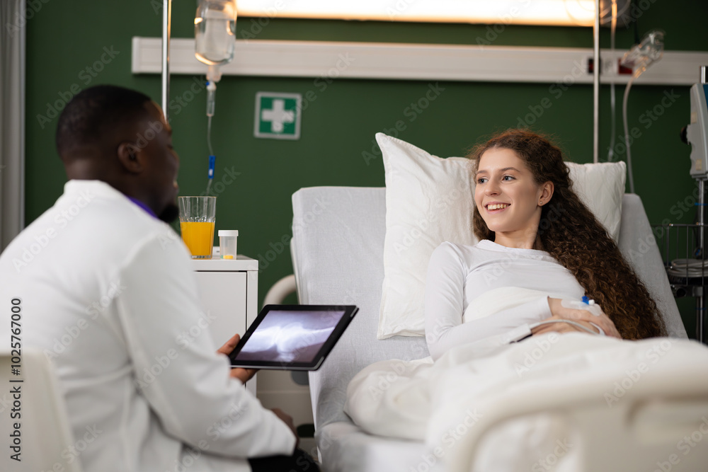 In the hospital room, the young woman, with a fracture, lies on the bed. Beside her, an attentive African doctor holds a tablet displaying an X-ray for a detailed consultation. In the hospital room, the young woman, with a fracture, lies on the bed. Beside her, an attentive African doctor holds a tablet displaying an X-ray for a detailed consultation.