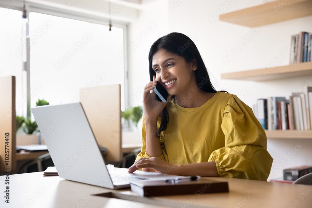 Happy busy young Indian student woman talking on cellphone in co-working, typing on laptop, using wireless technology for online communication, making work call, smiling, laughing Happy busy young Indian student woman talking on cellphone in co-working, typing on laptop, using wireless technology for online communication, making work call, smiling, laughing
