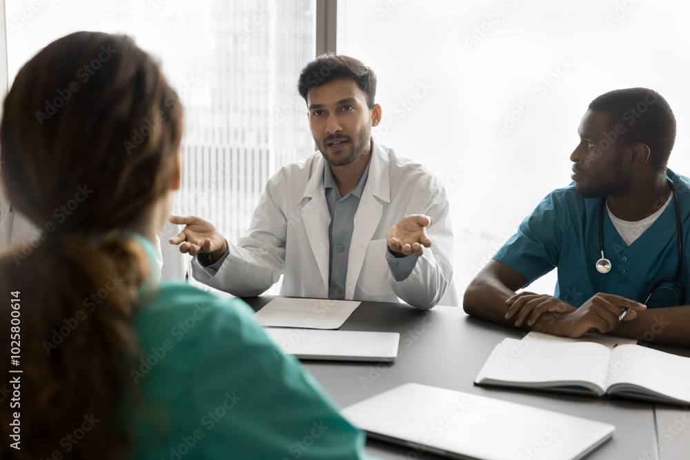 Young male doctor speak on multicultural team meeting ask advise in diagnostics choosing medical care tactics for severe patient case. Diverse medics colleagues meet for briefing at clinic staff room Young male doctor speak on multicultural team meeting ask advise in diagnostics choosing medical care tactics for severe patient case. Diverse medics colleagues meet for briefing at clinic staff room