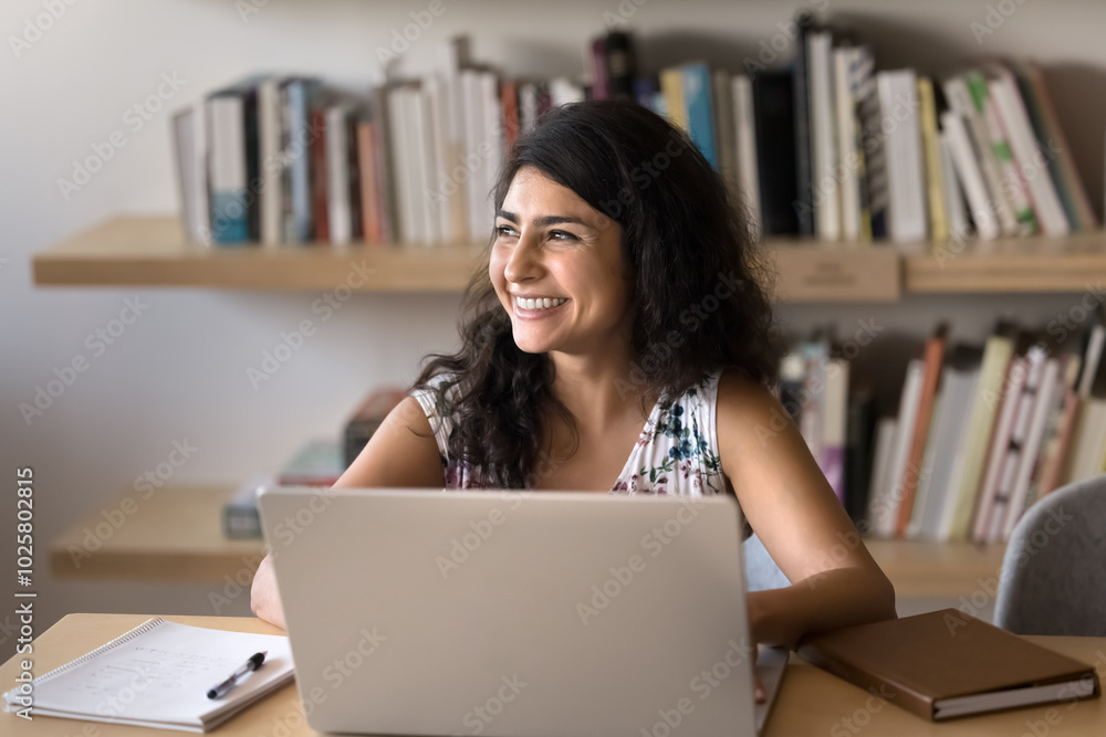 Happy young Indian project manager woman working on business project, doing research study in library, sitting at laptop with bookshelves in background, looking away, thinking, smiling, laughing Happy young Indian project manager woman working on business project, doing research study in library, sitting at laptop with bookshelves in background, looking away, thinking, smiling, laughing