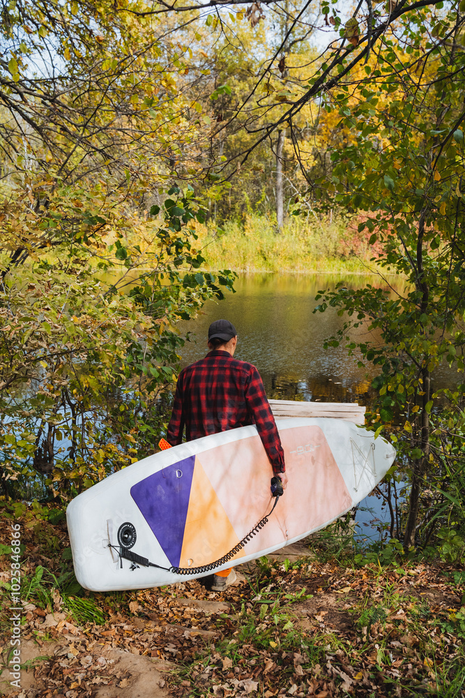 A man holds a SUP board in his hand and goes down to the river, SUP surfing on a reservoir, a solo trip across the lake on a paddle board. A man holds a SUP board in his hand and goes down to the river, SUP surfing on a reservoir, a solo trip across the lake on a paddle board.