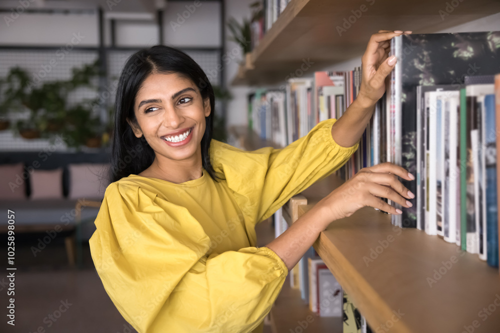 Fototapeta premium Cheerful young Indian student girl enjoying reading hobby, choosing book in library, bookstore, taking textbook from bookshelf, looking away, smiling, laughing, looking for information