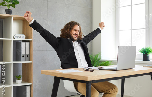 Relaxed satisfied business man stretching at the desk on workplace and looking on computer monitor screen. Young attractive confident bearded employee with long hair working on a laptop at office.