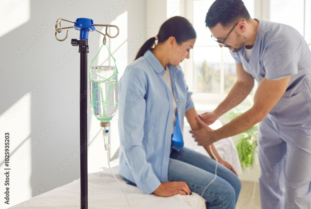 Fototapeta premium Nurse setting up IV line for patient. Close up glass drip bottle on stand in hospital ward, with male doctor inserting needle in woman's arm in background. Vitamin therapy, medical procedure concept