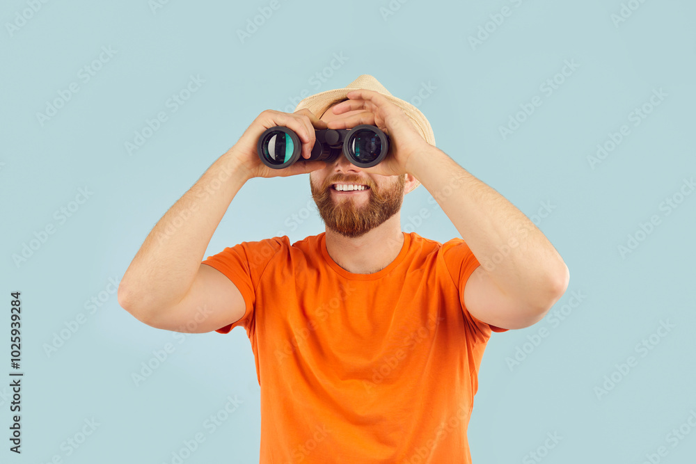 Portrait of happy smiling young man tourist wearing casual clothes and beach hat looking through binoculars on studio blue background. Summer holidays trip, search and vacation concept. Portrait of happy smiling young man tourist wearing casual clothes and beach hat looking through binoculars on studio blue background. Summer holidays trip, search and vacation concept.