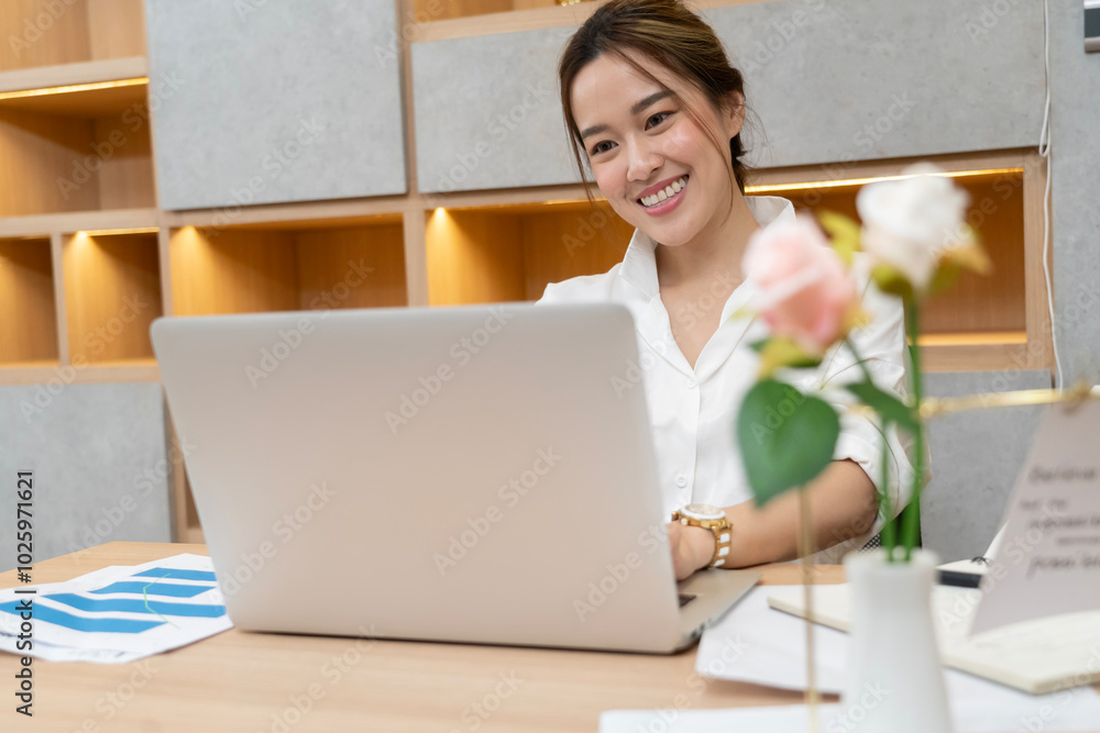 A woman is sitting at a desk with a laptop open in front of her. She is smiling and she is enjoying her work. The desk is cluttered with papers and books, and there is a potted plant in the background A woman is sitting at a desk with a laptop open in front of her. She is smiling and she is enjoying her work. The desk is cluttered with papers and books, and there is a potted plant in the background