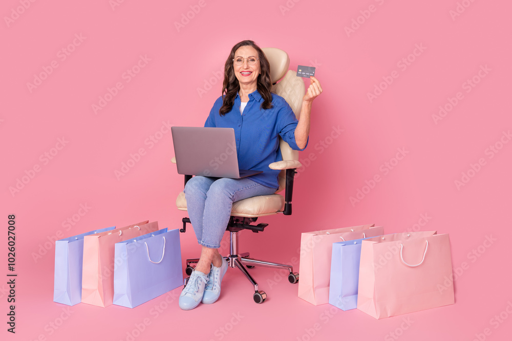 Full length photo of lovely senior lady sit armchair netbook pay shopper dressed stylish blue garment isolated on pink color background Full length photo of lovely senior lady sit armchair netbook pay shopper dressed stylish blue garment isolated on pink color background