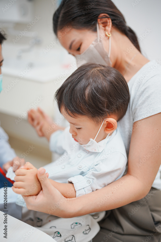 Mother and child wearing masks, sitting together in a healthcare setting, practicing safety measures during a medical visit. Mother and child wearing masks, sitting together in a healthcare setting, practicing safety measures during a medical visit.