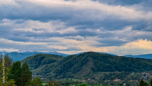 Breathtaking landscape featuring rolling hills covered in lush green forests near Graz, Styria. Majestic mountain range stands tall, with snow-capped peaks. Scene is one of natural beauty and serenity