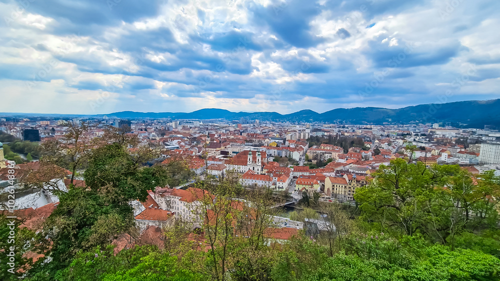 Fototapeta premium Tree branches framing scenic aerial view of Graz seen from Schlossberg, Styria, Austria, Europe. Unesco world heritage site with red-roofed buildings. City is nestled amidst lush green rolling hills
