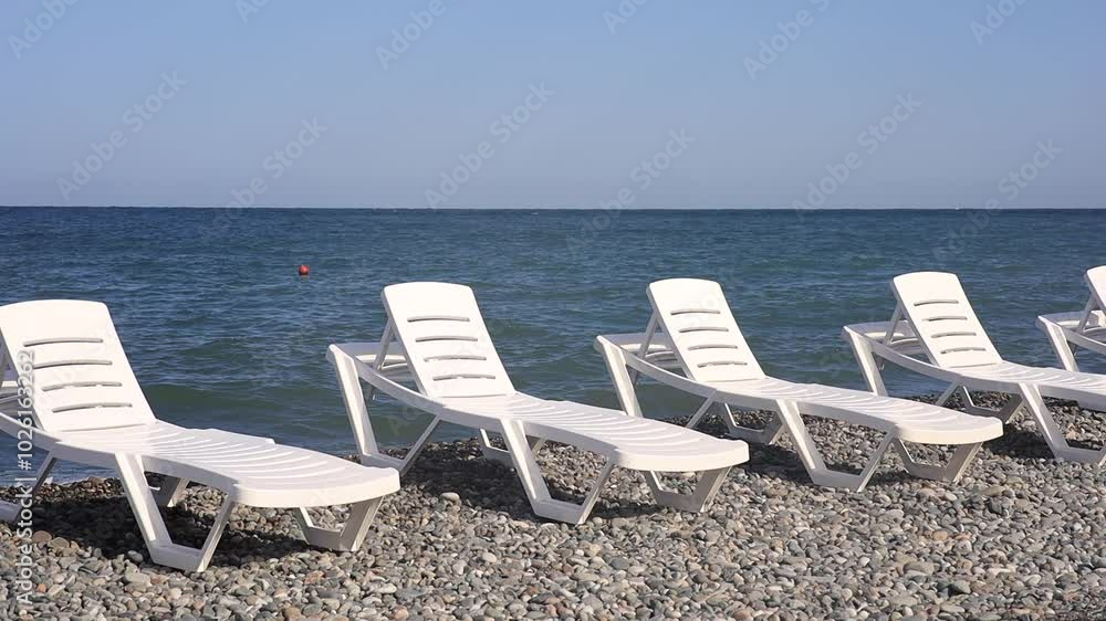 Empty white lounge chairs are arranged on a pebble beach by the calm blue sea, under a clear sky. The serene setting suggests a relaxing vacation spot, with no people in sight, perfect for unwinding.