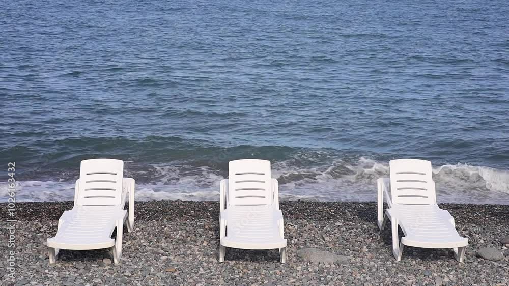 Empty white lounge chairs are arranged on a pebble beach by the calm blue sea, under a clear sky. The serene setting suggests a relaxing vacation spot, with no people in sight, perfect for unwinding. Empty white lounge chairs are arranged on a pebble beach by the calm blue sea, under a clear sky. The serene setting suggests a relaxing vacation spot, with no people in sight, perfect for unwinding.