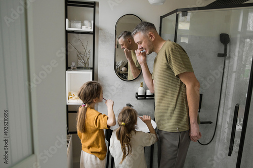 A father and his two daughters are all brushing their teeth in a modern bathroom. They are all looking in the mirror and smiling, making it a fun and healthy family activity.