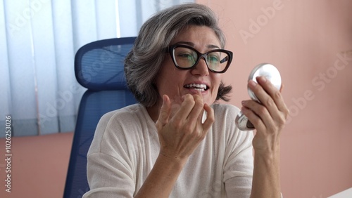 miling woman applies makeup, gray hair, glasses, wearing white top, holding mirror, sitting on blue chair, pink wall visible. Casual, relaxed moment reflecting confidence and ease.