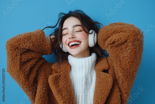 Young woman happily enjoying music while wearing cozy earmuffs against a blue...