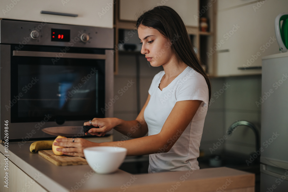 A focused young woman slices fruit on a wooden board in a stylish kitchen, demonstrating healthy meal preparation. She wears a white t-shirt, emphasizing a casual home cooking scene. A focused young woman slices fruit on a wooden board in a stylish kitchen, demonstrating healthy meal preparation. She wears a white t-shirt, emphasizing a casual home cooking scene.