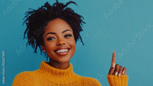 Cheerful woman in yellow sweater points upwards against a blue background in ...
