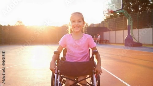 A young girl in a pink shirt is sitting in a wheelchair on a basketball court. She is doing her rehabilitation process and enjoying sport