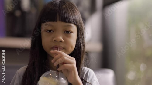 A cute Thai girl is drinking water from a brown straw. After tasting it, she makes a funny face because the drink in her glass has too much lemon sourness.