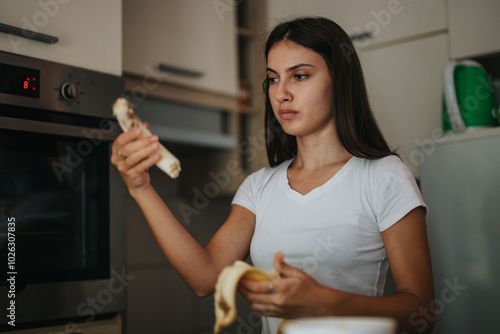 A focused young woman examines a peeled banana in her modern kitchen. The scene suggests contemplation and a possible decision-making moment in her daily routine.