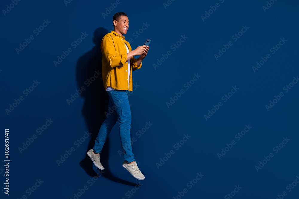 Full body length photo of young guy wear jeans yellow shirt jumping with smartphone going smiling isolated on dark blue color background Full body length photo of young guy wear jeans yellow shirt jumping with smartphone going smiling isolated on dark blue color background