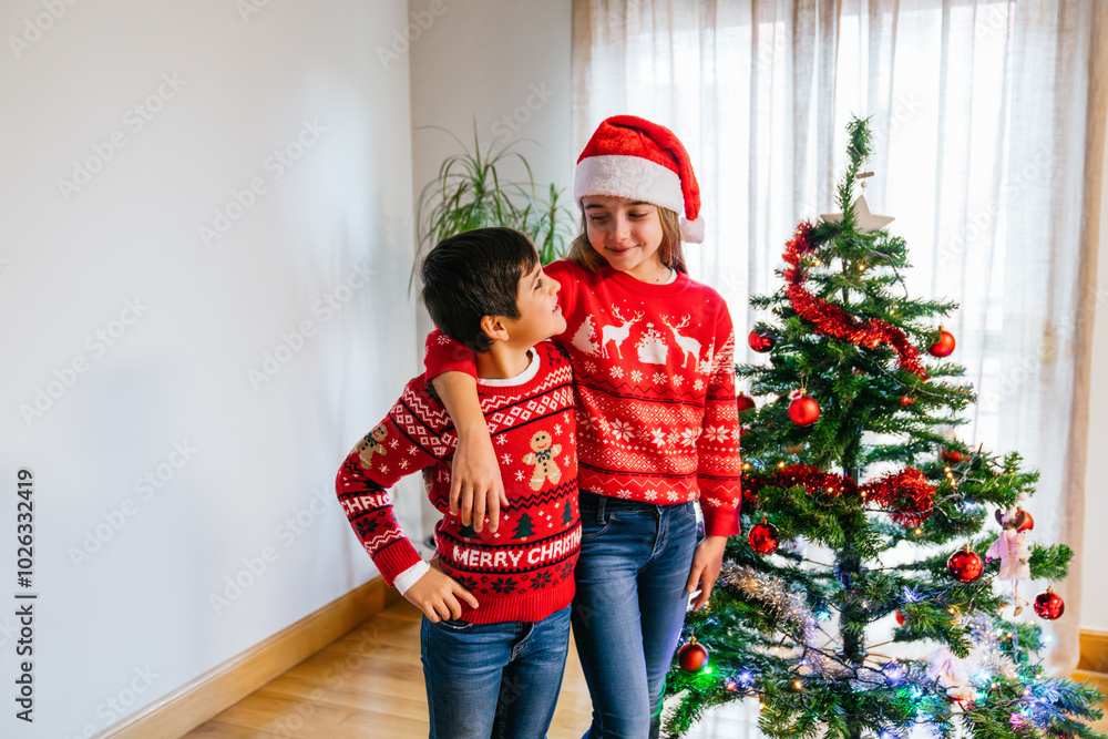 Siblings posing proudly beside their Christmas tree in cozy festive outfits Siblings posing proudly beside their Christmas tree in cozy festive outfits