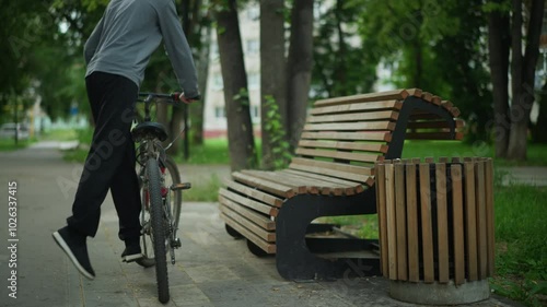 Young boy with glasses in gray top steps off bicycle near wooden bench in park, adjusting bicycle close to bench before sitting down and scratching face, with trees, greenery, and urban background
