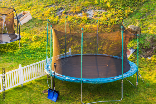 Trampoline with safety net in backyard, with snow shovel leaning against it, set against grassy slope in background. Sweden. photo