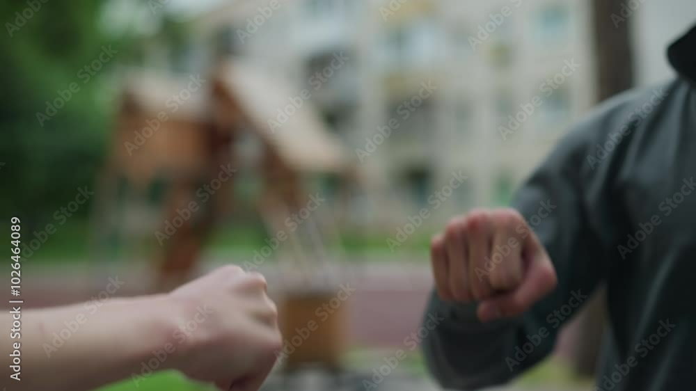Close-up of hands interacting in a playful knuckle bump, one person in a grey top extends their hand to connect with another, with a blurred background featuring residential buildings Close-up of hands interacting in a playful knuckle bump, one person in a grey top extends their hand to connect with another, with a blurred background featuring residential buildings