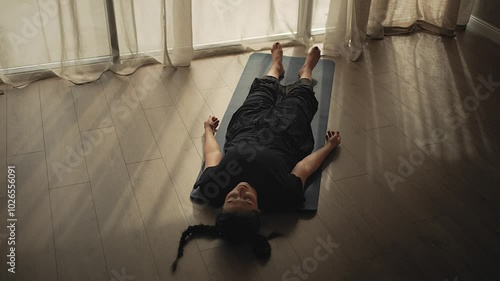 Top shot of a young woman laying down on the yoga mat, relaxing and inhaling after the yoga class. Light rays on the wooden floor.
