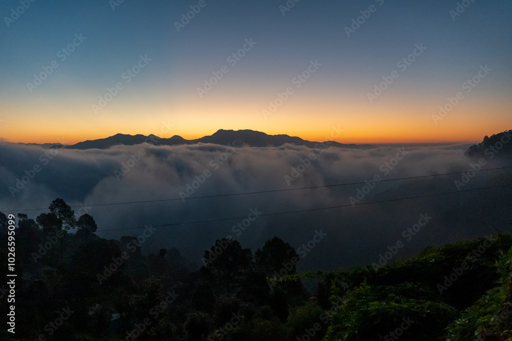 Before sunrise view, painted sky with soft orange light in the horizon. Dense clouds covers the valley and mountains in background above the clouds. Magical morning scene in mountain village in India. Before sunrise view, painted sky with soft orange light in the horizon. Dense clouds covers the valley and mountains in background above the clouds. Magical morning scene in mountain village in India.